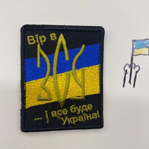 Puede incluir: Un parche bordado negro con el escudo de armas ucraniano en amarillo y azul. El parche tiene las palabras "Вір в" en la parte superior y "- і все буде Україна!" en la parte inferior. A la derecha hay un pequeño dibujo de una bandera ucraniana.