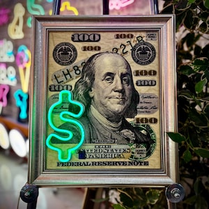 May include: A framed print of a US $100 bill with a green neon dollar sign in front of it. The bill features a portrait of Benjamin Franklin and the words "Federal Reserve Note".