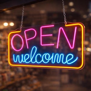 Könnte beinhalten: Ein Neonschild mit den Worten "OPEN welcome" in Pink und Blau, umrandet von Gelb, hängt an Ketten. Der unscharfe Hintergrund deutet auf ein Geschäft mit warmer Beleuchtung hin.