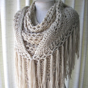 May include: A cream-colored knitted shawl with a delicate lace pattern and long fringe. The shawl is draped over a mannequin.