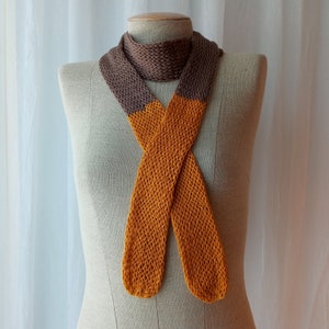 May include: A brown and orange knitted scarf with a criss-cross pattern. The scarf is displayed on a mannequin.
