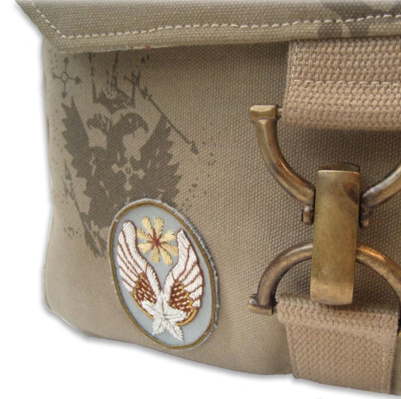 Peut inclure: Gros plan sur un sac en toile beige avec un &eacute;cusson d&eacute;coratif et une garniture m&eacute;tallique. L'&eacute;cusson pr&eacute;sente un motif brod&eacute; d'ailes et d'une fleur. Le sac a un motif d'aigle vieilli et un fermoir couleur laiton.