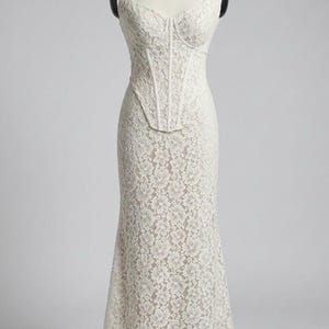 May include: A white lace gown displayed on a mannequin. The dress features a fitted bodice with a corset-style design and a sweetheart neckline. The skirt flows into a subtle mermaid silhouette. The entire dress is covered in a delicate floral lace pattern.