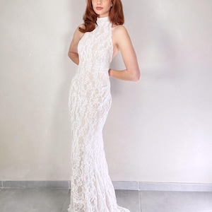 May include: A white lace gown with a high neckline and open back. The floor-length dress features a fitted silhouette that flares out at the hem. The intricate lace detailing covers the entire dress, creating a delicate and elegant look. The model is standing in front of a plain white wall.