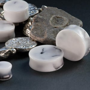 May include: White and black marbled ear plugs in various sizes are displayed. The plugs have a smooth, polished finish and a rounded shape. A gray rock and silver-colored metal pieces are also in the image, set against a dark background.