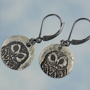 May include: Silver owl earrings with a detailed etched design. The earrings are on a silver hook.