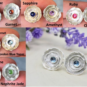 May include: A collection of sterling silver flower-shaped stud earrings. Each pair features a different gemstone center, including white topaz, garnet, sapphire, ruby, amethyst, citrine, rhodolite garnet, London blue topaz, tourmaline, and nephrite jade.