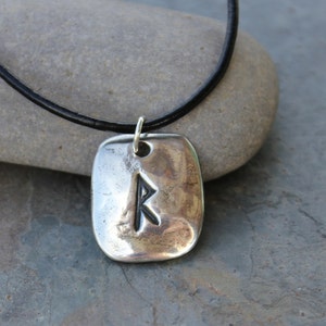 May include: A silver-toned pendant with a black leather cord. The pendant has a rectangular shape with rounded edges and a carved rune symbol. The necklace is displayed on a gray stone surface.