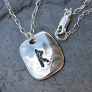 May include: A silver pendant necklace featuring a rectangular, textured pendant with a black rune symbol. The pendant hangs from a delicate silver chain with a lobster clasp. The necklace is displayed on a dark, textured surface.