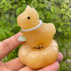 May include: A light yellow cat figurine with a pearl necklace and a small green charm. The cat is sitting on a round base. The figurine is made of a translucent material.
