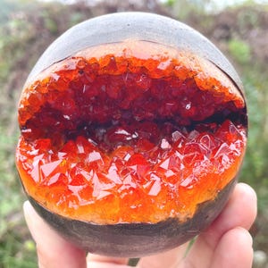 May include: A spherical geode with a dark exterior and a vibrant interior. The inside is filled with sparkling, deep red and orange crystals. The geode is held in front of a blurred green background, showcasing its natural beauty and unique color.