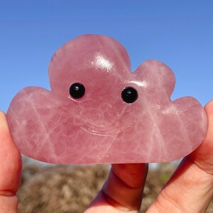 May include: A pink rose quartz cloud-shaped carving with black eyes and a smiling mouth. The gemstone is held up against a clear blue sky. The cloud carving is a unique decorative item.
