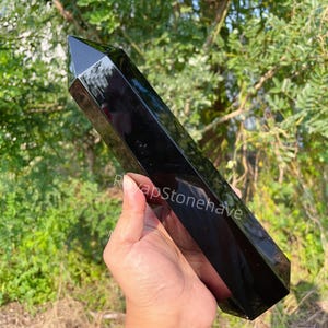 May include: A polished black obsidian obelisk, held against a backdrop of green foliage. The crystal has a smooth, reflective surface and a pointed top. The obelisk is a dark, solid color, and appears to be a natural stone.