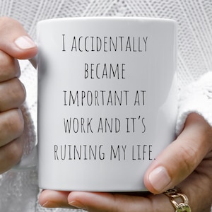 Op de afbeelding: Een witte keramische mok met de tekst "I ACCIDENTALLY BECAME IMPORTANT AT WORK AND IT'S RUINING MY LIFE" in het zwart. De mok wordt vastgehouden door een persoon met gouden ringen aan hun vingers.