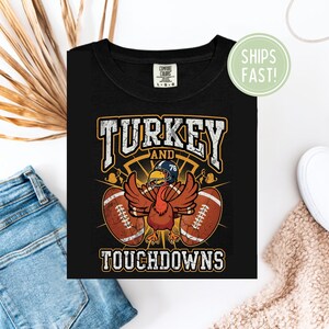 Könnte beinhalten: Schwarzes T-Shirt mit einer Grafik einer Truthahn mit Footballhelm, ausgebreiteten Flügeln und zwei Fußbällen. Der Text lautet "TURKEY AND TOUCHDOWNS" in einem kräftigen Vintage-Stil. Das Shirt ist von der Marke Comfort Colors.
