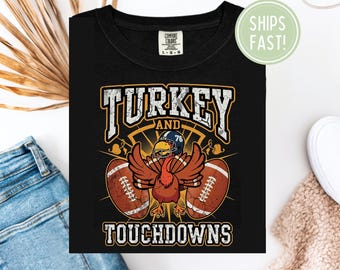 Puede incluir: Camiseta negra con un diseño de un pavo con casco de fútbol, alas extendidas y dos balones de fútbol. El texto dice "TURKEY AND TOUCHDOWNS" en un estilo vintage audaz. La camiseta es de la marca Comfort Colors.