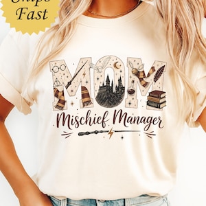 Op de afbeelding: Een crèmekleurig t-shirt met het woord MOM in grote blokletters gevuld met magische illustraties, waaronder een kasteel, een uil, een toverstaf en boeken. Onder het woord MOM staat de tekst Mischief Manager in een donkerbruin lettertype.