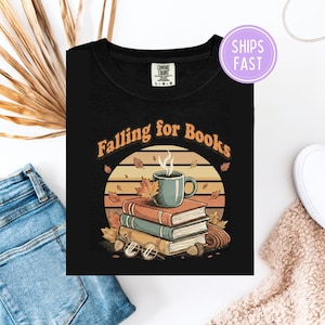May include: Black t-shirt with a graphic featuring a stack of books, a coffee mug, and autumn leaves. The text "Falling for Books" is arched above the design. The shirt has a crew neck and short sleeves.