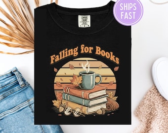 May include: Black t-shirt with a graphic featuring a stack of books, a coffee mug, and autumn leaves. The text "Falling for Books" is arched above the design. The shirt has a crew neck and short sleeves.