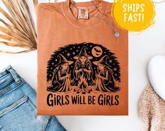 May include: An orange t-shirt with a black graphic of three witches around a campfire, with the text "GIRLS WILL BE GIRLS". A silver necklace is draped over the shirt. The shirt is folded on a white surface with blue jeans and a beige scarf.