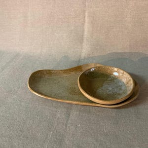 Peut inclure: Un ensemble de service en céramique fait à la main, comprenant un plateau long et de forme irrégulière et un petit bol rond. L'ensemble présente un glaçage vert et brun moucheté, idéal pour les apéritifs.