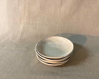 May include: A stack of four white ceramic bowls with a speckled glaze. The bowls are round and shallow, and they are stacked on top of each other.