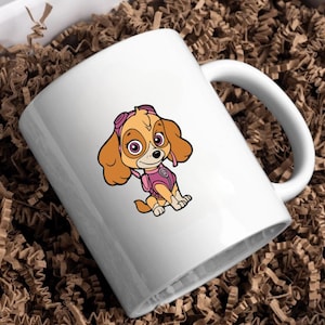 Puede incluir: Taza de cer&aacute;mica blanca con una ilustraci&oacute;n de dibujos animados de Skye de Paw Patrol. El personaje tiene pelaje marr&oacute;n claro, gafas rosas y un chaleco rosa. La taza tiene un asa curva y est&aacute; sobre papel de embalaje marr&oacute;n.