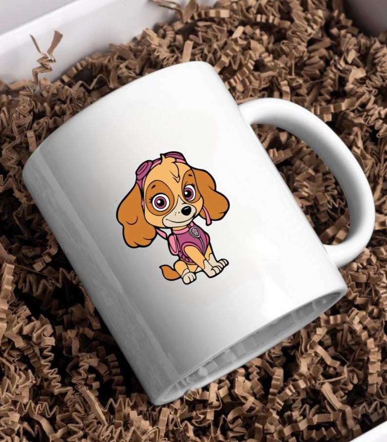 Puede incluir: Taza de cer&aacute;mica blanca con una ilustraci&oacute;n de dibujos animados de Skye de Paw Patrol. El personaje tiene pelaje marr&oacute;n claro, gafas rosas y un chaleco rosa. La taza tiene un asa curva y est&aacute; sobre papel de embalaje marr&oacute;n.