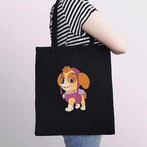 Puede incluir: Bolso tote negro con una ilustraci&oacute;n de dibujos animados de Skye de Paw Patrol. El personaje est&aacute; representado en tonos rosa, naranja y beige. El bolso tiene asas negras y lo lleva una persona con una camiseta a rayas blancas y negras.