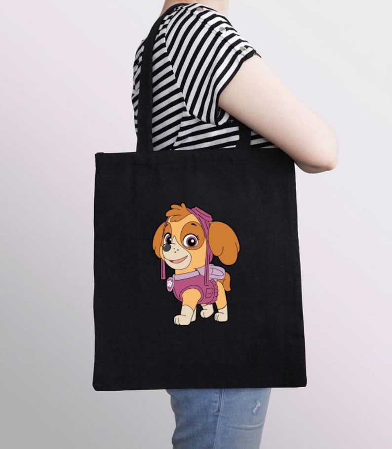 Puede incluir: Bolso tote negro con una ilustraci&oacute;n de dibujos animados de Skye de Paw Patrol. El personaje est&aacute; representado en tonos rosa, naranja y beige. El bolso tiene asas negras y lo lleva una persona con una camiseta a rayas blancas y negras.