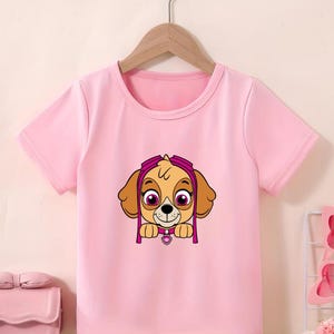 Puede incluir: Camiseta rosa claro de manga corta con un dibujo animado de un cachorro marr&oacute;n con gafas rosas y un colgante en forma de coraz&oacute;n rosa. El cachorro tiene orejas marrones y ojos grandes. La camiseta est&aacute; colgada de una percha de madera.