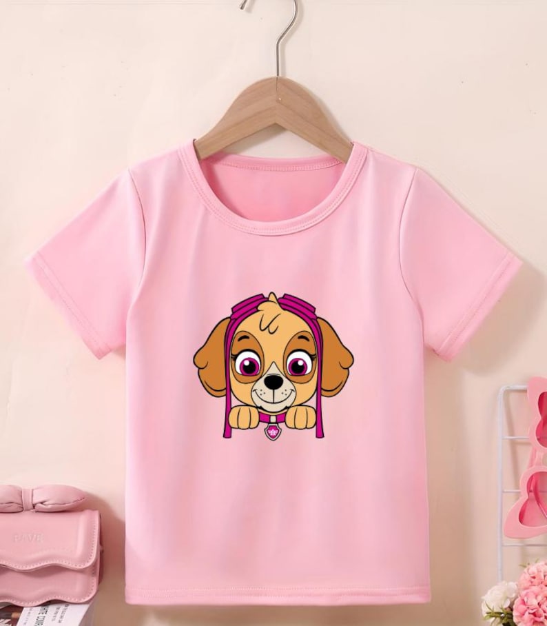 Puede incluir: Camiseta rosa claro de manga corta con un dibujo animado de un cachorro marr&oacute;n con gafas rosas y un colgante en forma de coraz&oacute;n rosa. El cachorro tiene orejas marrones y ojos grandes. La camiseta est&aacute; colgada de una percha de madera.