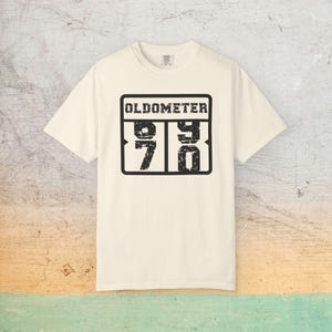 May include: Cream-colored t-shirt with a black graphic that reads "OLDOMETER" above the numbers "98" and "70" inside a black rectangle. The shirt has short sleeves and a crew neck.
