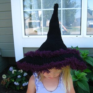 May include: A black crochet witch hat with a wide brim and a purple fringe. The hat has a tall, pointed crown and is likely handmade. The hat is a Halloween costume accessory.