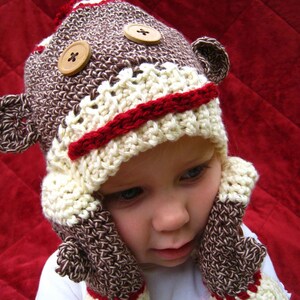 May include: A handmade crochet monkey hat and matching mittens set. The hat is brown, cream, and red with wooden button accents and ear details. The mittens have a similar color scheme.