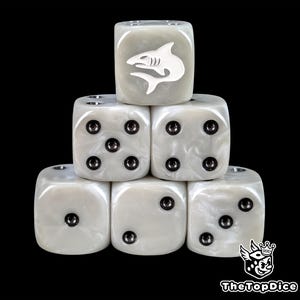 May include: Six pearlescent white dice are stacked. One die has a white shark graphic. The dice have black pips. The dice are arranged in a pyramid shape against a black background. The dice are for tabletop games.