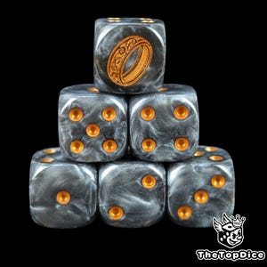 May include: A stack of six dice with a dark gray, marbled appearance and gold-colored pips. One die features a gold-colored ring design. The dice are for tabletop games and are set against a black background.