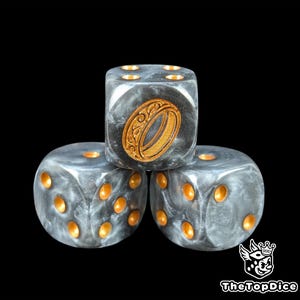 May include: Three gray and gold dice with a black background. The dice have gold pips and a gold ring design on one face. The dice are stacked on top of each other.
