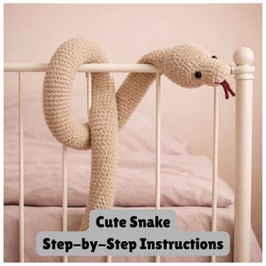 Peut inclure: Un serpent en crochet beige drapé sur un cadre de lit en métal blanc. Le serpent a des yeux noirs et une langue fourchue rouge. Le texte "Cute Snake Step-by-Step Instructions" est au bas de l'image.