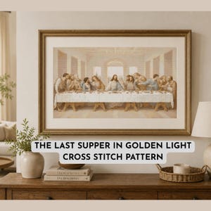 Op de afbeelding: Een ingelijst kruissteekpatroon getiteld The Last Supper in Golden Light, met een groep figuren aan een lange tafel in een neutraal, sepia-kleurenpalet, gepresenteerd in een goudkleurige lijst op een houten ondergrond.