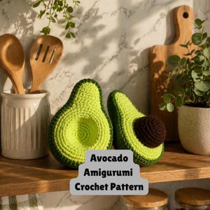 May include: Two crocheted avocado halves with a dark brown pit sit on a wooden shelf. The avocados are green with a darker green border. The text "Avocado Amigurumi Crochet Pattern" is displayed on a gray background.
