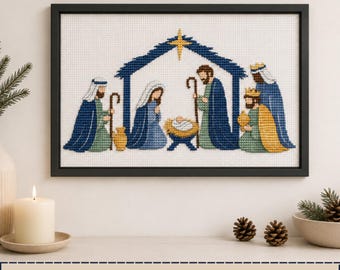 Op de afbeelding: Een ingelijst kruissteek kunstwerk met een kerststalscène, inclusief een centrale stalstructuur, een gele ster en figuren zoals Maria, Jozef en drie koningen rondom een kribbe, uitgevoerd in blauwe, groene en gouden draadkleuren.