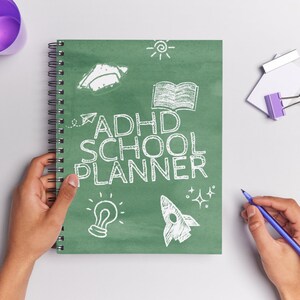 Może przedstawiać: Spiralny notatnik z zieloną okładką z białym napisem "ADHD SCHOOL PLANNER". Okładka zawiera r&oacute;wnież białe ilustracje żar&oacute;wki, rakiety, książki, słońca i samolotu. Ręka trzyma notatnik, a druga ręka trzyma niebieski długopis.