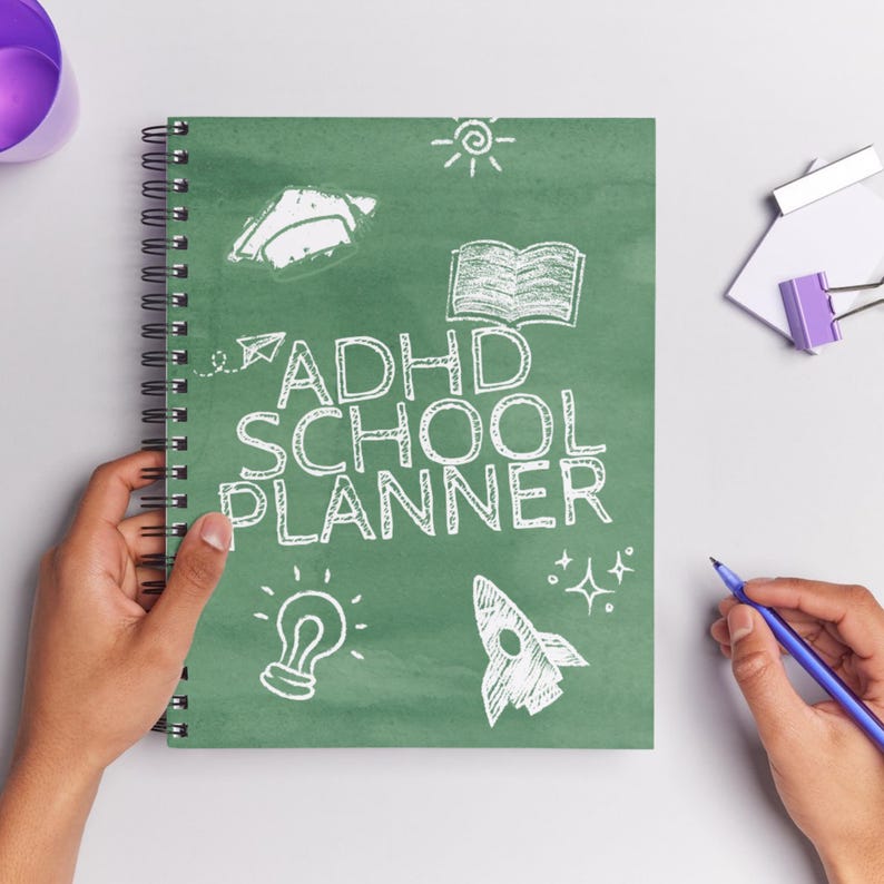 Może przedstawiać: Spiralny notatnik z zieloną okładką z białym napisem "ADHD SCHOOL PLANNER". Okładka zawiera r&oacute;wnież białe ilustracje żar&oacute;wki, rakiety, książki, słońca i samolotu. Ręka trzyma notatnik, a druga ręka trzyma niebieski długopis.
