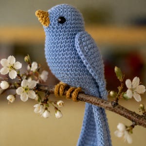 May include: A handmade blue crochet bird with a yellow beak and feet perches on a brown branch with white blossoms. The bird has a black eye and is made of soft yarn. A charming, decorative item.