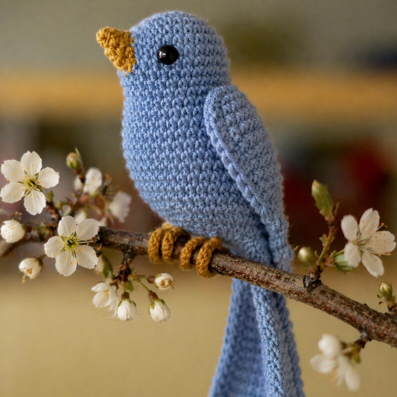 May include: A handmade blue crochet bird with a yellow beak and feet perches on a brown branch with white blossoms. The bird has a black eye and is made of soft yarn. A charming, decorative item.