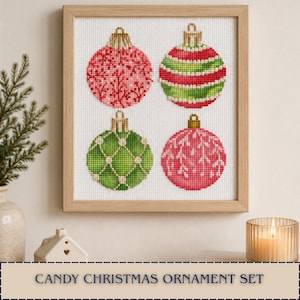 May include: A square wooden frame containing a cross-stitch embroidery of four Christmas ornaments. The ornaments feature pink and green colors with intricate patterns, including stripes, grids, and branch-like designs, all set against a white fabric.