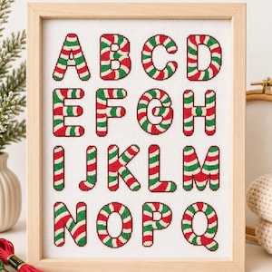 May include: A framed cross-stitch embroidery on white fabric featuring the letters A through Q. Each letter is rendered in a candy cane pattern with diagonal red, white, and green stripes. The artwork is enclosed in a light-colored wooden frame.