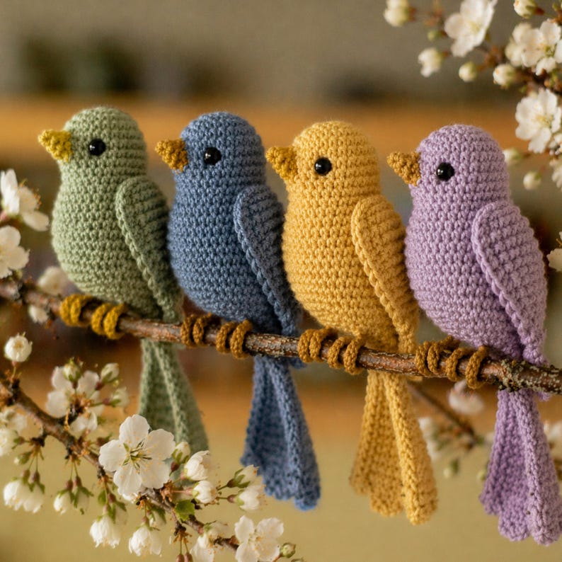 May include: Four crocheted birds in shades of green, blue, yellow, and purple perch on a brown branch. Each bird has a contrasting yellow beak and black eyes. The branch is adorned with brown yarn and set against a backdrop of white flowers.