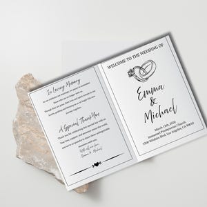 Può includere: Un programma di nozze bianco con testo nero. La copertina anteriore presenta una semplice illustrazione di due anelli intrecciati e i nomi "Emma & Michael". La copertina posteriore include un messaggio di ringraziamento agli ospiti e una nota in memoria amorevole di coloro che sono scomparsi.